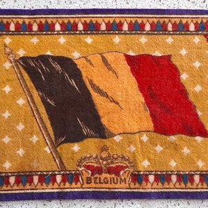 May include: A vintage woven tapestry featuring the Belgian flag with a crown and the word "BELGIUM" at the bottom. The tapestry has a gold background with a white diamond pattern and a colorful border.