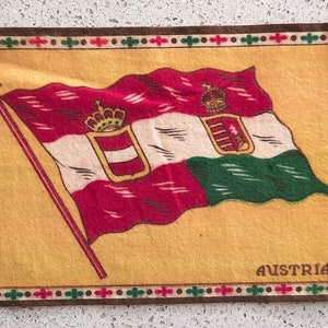 May include: A vintage matchbook cover featuring a red, white, and green flag with a coat of arms and the word "AUSTRIA" printed below. The flag is on a yellow background with a brown border.