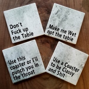 May include: Four square coasters with humorous sayings printed on them. The coasters are made of a light gray stone-like material with a rough texture. The sayings are: "Don't Fuck up the Table", "Make me Wet not the Table", "Use this coaster or I'll punch you in the throat", and "Use a Coaster to be Classy and Shit".