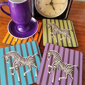 May include: Four square coasters with a zebra design. Each coaster has a different colored background: orange, green, blue, and purple. The zebra is black and white with a striped pattern.