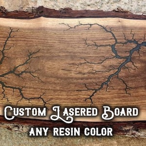 May include: A custom-made wooden board with a live edge, featuring a unique laser-etched design resembling lightning. The dark resin-filled cracks create a striking contrast against the wood grain. The text "CUSTOM LASERED BOARD ANY RESIN COLOR" is displayed.