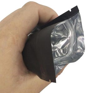May include: A small, rectangular stand-up pouch featuring a matte black exterior and a metallic silver interior lining. The pouch includes a clear plastic window on one side and a black heat-sealed top edge with a small notch for easy opening.