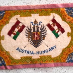 May include: A vintage fabric patch with a pink border and a white center. The patch features a coat of arms with a double-headed eagle and two crossed flags. The flags are red, white, and green. The text "AUSTRIA-HUNGARY" is printed in blue below the coat of arms.