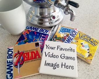 May include: Four different video game cartridges on coasters. The cartridges are for the Game Boy, Game Boy Advance, and Pokemon. The coasters are white with black text that says "Your Favorite Video Game Image Here".