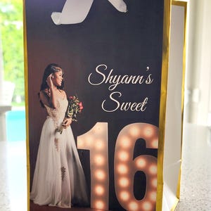 K&ouml;nnte beinhalten: Eine goldene Geschenkt&uuml;te mit einem wei&szlig;en Band. Die Tasche zeigt ein Foto einer jungen Frau in einem wei&szlig;en Kleid, die einen Blumenstrau&szlig; h&auml;lt. Der Text "Shyann's Sweet 16" ist auf der Tasche aufgedruckt.