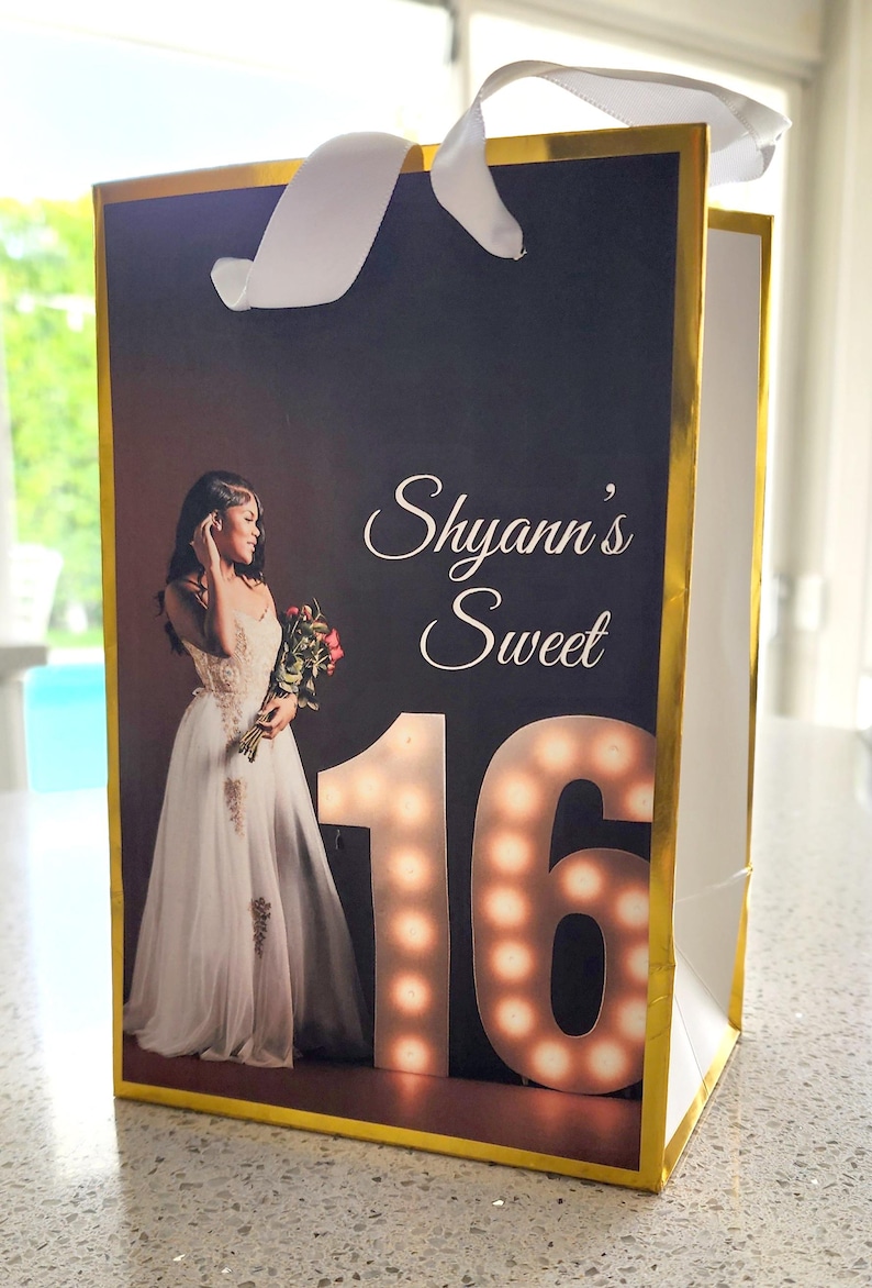 K&ouml;nnte beinhalten: Eine goldene Geschenkt&uuml;te mit einem wei&szlig;en Band. Die Tasche zeigt ein Foto einer jungen Frau in einem wei&szlig;en Kleid, die einen Blumenstrau&szlig; h&auml;lt. Der Text "Shyann's Sweet 16" ist auf der Tasche aufgedruckt.