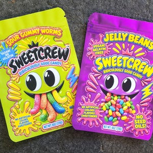 May include: Two stand-up pouches of Sweetcrew candy. The lime green pouch features Sour Gummy Worms, while the purple pouch features Jelly Beans. Both bags display a cartoon character, the text Ridiculously Good Candy, and net weight 6oz (170g).