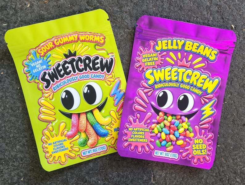 May include: Two stand-up pouches of Sweetcrew candy. The lime green pouch features Sour Gummy Worms, while the purple pouch features Jelly Beans. Both bags display a cartoon character, the text Ridiculously Good Candy, and net weight 6oz (170g).