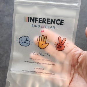 May include: A translucent, resealable coffee pouch labeled INFERENCE BIRD and BEAR. The front features rock, paper, scissors hand icons, the text Ro-Sham-Bo, and flavor notes of mulling spice, plum, and dark chocolate. It contains 4 oz of coffee.