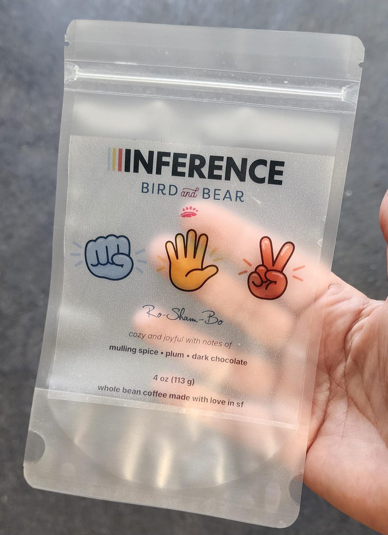 May include: A translucent, resealable coffee pouch labeled INFERENCE BIRD and BEAR. The front features rock, paper, scissors hand icons, the text Ro-Sham-Bo, and flavor notes of mulling spice, plum, and dark chocolate. It contains 4 oz of coffee.