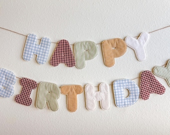 May include: A fabric banner spelling "HAPPY BIRTHDAY" in various gingham and striped patterns. The letters are in blue, red, green, yellow, and white, strung together with twine, perfect for a birthday celebration.