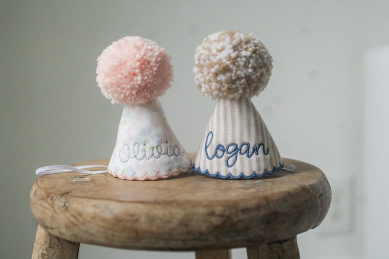 May include: Two personalized party hats with pom-poms. One hat is pink and floral, embroidered with "Olivia." The other is beige striped, embroidered with "Logan." Both have scalloped edges and are on a wooden stool.