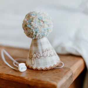May include: A whimsical, handmade party hat with a pom-pom top in pastel colors. The hat is made of a beige and white checkered fabric, with the word "poetry" embroidered in a cursive font. A white elastic cord is attached.