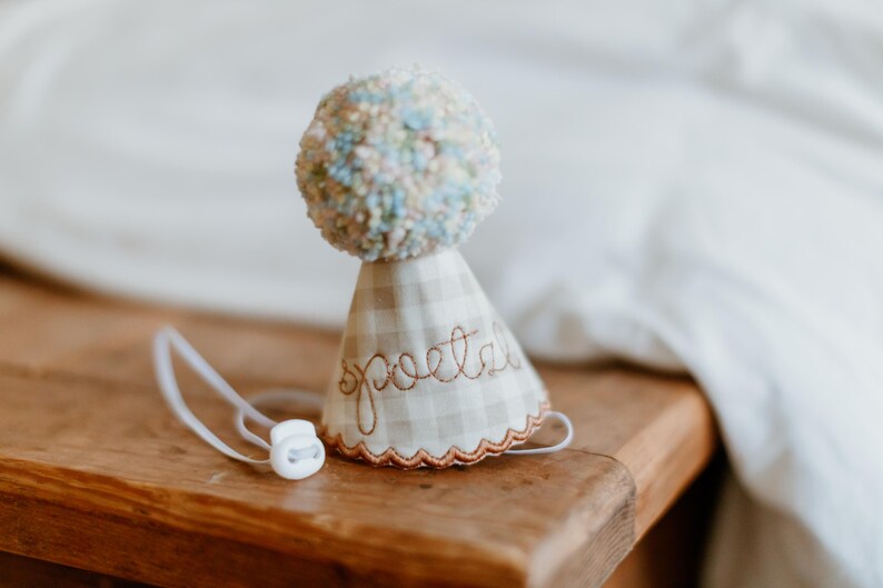 May include: A whimsical, handmade party hat with a pom-pom top in pastel colors. The hat is made of a beige and white checkered fabric, with the word "poetry" embroidered in a cursive font. A white elastic cord is attached.