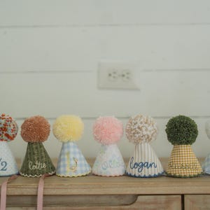May include: A row of colorful party hats with pom-poms. Each hat has a unique fabric pattern and is personalized with names or phrases like "Happy Birthday Tchal" and "1/2". The hats are displayed on a wooden surface.