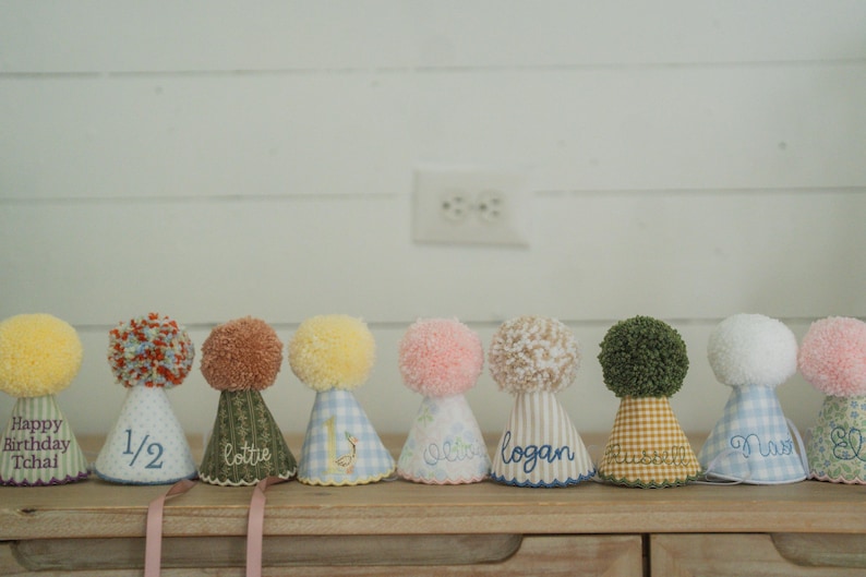 May include: A row of colorful party hats with pom-poms. Each hat has a unique fabric pattern and is personalized with names or phrases like "Happy Birthday Tchal" and "1/2". The hats are displayed on a wooden surface.