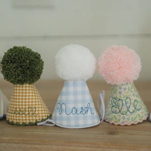 May include: A row of personalized birthday party hats with pom-poms. The hats feature various patterns, including stripes, gingham, and floral prints. Each hat is embroidered with a name or phrase, such as "Logan," "Russell," and "Birthday Boy."