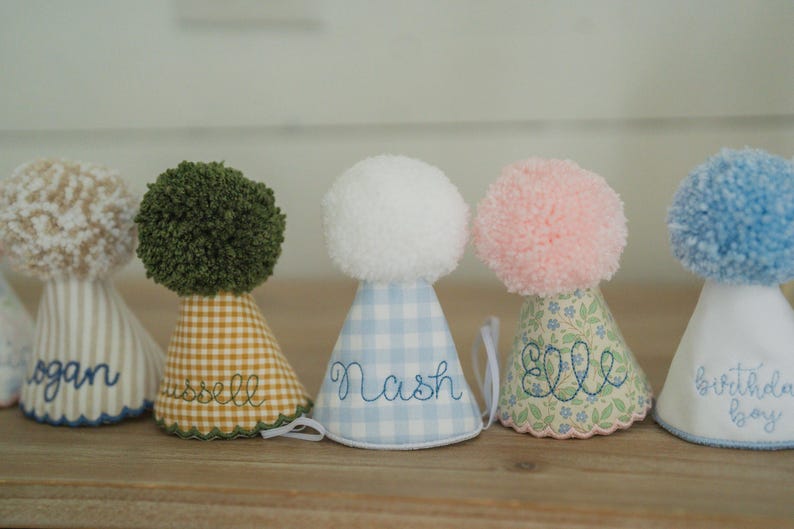 May include: A row of personalized birthday party hats with pom-poms. The hats feature various patterns, including stripes, gingham, and floral prints. Each hat is embroidered with a name or phrase, such as "Logan," "Russell," and "Birthday Boy."