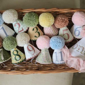 Könnte beinhalten: Eine Sammlung bunter Geburtstagshüte mit Pompons und den Zahlen 1-11 sowie einem "one"-Hut, angeordnet in einem Weidenkorb. Die Hüte weisen verschiedene Muster und Farben auf, darunter Blau, Grün, Rosa und Gelb. Der Korb ist aus Weide geflochten.