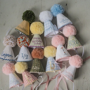 May include: A collection of handmade party hats in various colors and patterns. Each hat is adorned with a fluffy pom-pom and personalized with names or numbers. The hats are made of fabric and have ribbons attached for securing.