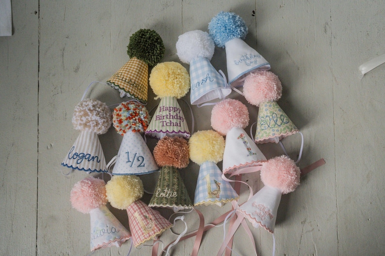 May include: A collection of handmade party hats in various colors and patterns. Each hat is adorned with a fluffy pom-pom and personalized with names or numbers. The hats are made of fabric and have ribbons attached for securing.
