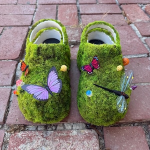 Op de afbeelding: Een paar groene, met mos bedekte klompen versierd met kleurrijke vlinders, paddenstoelen en een libelle. De klompen hebben witte bandjes en zwarte binnenzolen, wat een grillig, door de natuur geïnspireerd ontwerp creëert.