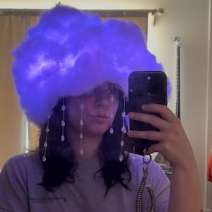 Puede incluir: Un tocado en forma de nube esponjosa, iluminado con un brillo púrpura. Gotas de cristal transparente cuelgan del borde de la nube. Este inusual tocado es un llamativo accesorio de disfraz, ideal para eventos temáticos.