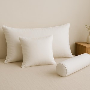 May include: A bed with three white pillows and a bolster. The pillows are rectangular and square, and the bolster is cylindrical. The bed is covered with a light beige sheet. A small wooden nightstand with a vase of flowers is in the background.