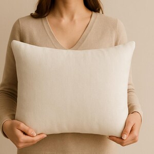 Puede incluir: Una almohada rectangular de color blanquecino se sostiene frente a una persona. La almohada tiene un color suave y neutro y un diseño sencillo. La almohada es un artículo de decoración del hogar, probablemente para una cama o un sofá.