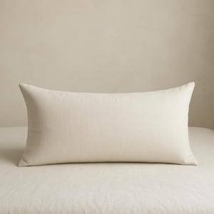 Puede incluir: Una almohada rectangular de color crema con una apariencia suave y texturizada. La almohada se coloca sobre una sábana de cama crema a juego, creando una estética cohesiva y neutra. Su forma sugiere que está diseñada para soporte lumbar o uso decorativo.