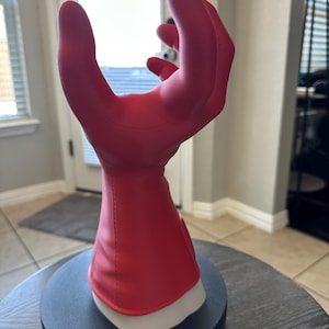 Può includere: Un guanto rosso brillante, forse per la pulizia o per uso industriale, è esposto su un supporto rotante nero. Il guanto è realizzato in un materiale liscio e ha un polsino interno bianco. Le dita sono curve verso l'alto.