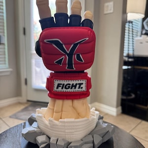 May include: A sculpted figurine of a hand wearing a red and black MMA-style glove with the text FIGHT on the wrist strap and a stylized Y logo on the back of the hand. The hand emerges from a white cracked base surrounded by gray rock-like debris.