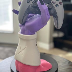 May include: A purple hand-shaped gaming controller holder with black fingernails, holding a gray and black controller. The holder has a white sleeve and a pink base, set on a black circular stand. A unique accessory for gamers.