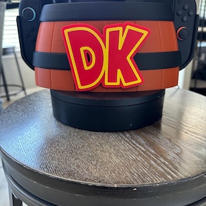Puede incluir: Una consola Nintendo Switch con una funda personalizada en forma de barril. La funda es marrón con bandas negras y presenta las letras "DK" en rojo con contornos amarillos. La consola está sobre un soporte giratorio negro.