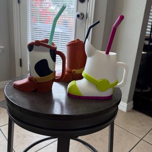 May include: Three boot-shaped mugs with handles and straws. One mug features a white and black cow-print pattern with a brown base. Another is solid brown with a cactus design. The third is white with lime green and purple accents.