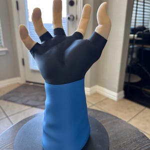 Puede incluir: Una escultura de una mano con un guante negro y una manga azul. Los dedos son de color carne y la mano está abierta. La escultura está montada sobre una base circular negra.