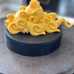 Könnte beinhalten: Eine dekorative Skulptur mit einem gelben Wolkenmuster auf einem schwarzen, runden Sockel. Die Wolke hat ein wirbelndes, strukturiertes Aussehen. Die Skulptur steht auf einem Holztisch.