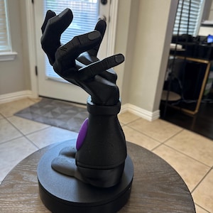 May include: A black sculpture of a hand reaching upwards, with a purple accent on a black base. The sculpture is on a round, dark wooden table. The hand is detailed and appears to be made of a solid material.