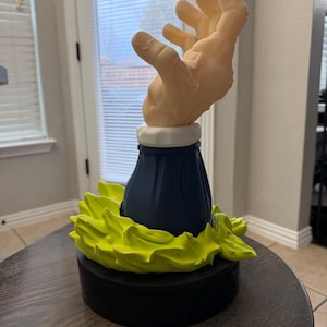 May include: A sculpture featuring a stylized hand emerging from a dark blue base with a white collar. The base is surrounded by bright yellow-green, flame-like shapes, all resting on a black circular platform. The sculpture is likely a decorative item.