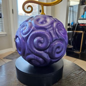 Peut inclure: Objet sphérique violet avec un motif tourbillonnant, surmonté d'une tige et d'une boucle dorées, posé sur une base noire. La surface est texturée, possiblement un objet décoratif.