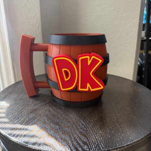 Puede incluir: Una taza marrón en forma de barril con un asa roja y las letras "DK" en rojo con un contorno amarillo. La taza tiene bandas negras alrededor del barril y está sobre una superficie de madera oscura.