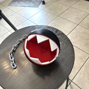 May include: A black and red decorative ball with white jagged edges, resembling a cartoon character. A silver chain is attached to the ball. The ball is sitting on a dark brown wooden stool.