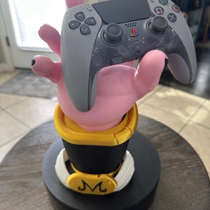 May include: A pink hand-shaped controller holder with a gray PlayStation 5 controller. The holder has a black base with a yellow and white trim, and a gold emblem. The controller has the standard button layout.