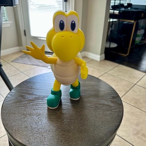 May include: A yellow Koopa Troopa figurine with a beige shell, green boots, and a friendly pose. The figure is standing on a dark brown stool. The figurine is a character from the Super Mario video game series.