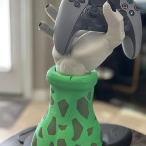 May include: A gray and black PlayStation 5 controller rests in a white hand-shaped holder. The holder is mounted on a green base with brown, irregular patterns. The base sits on a black, rounded platform.