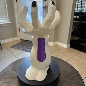 May include: A white sculpture of a hand with black fingernails, holding an open position. The base is white with a purple accent and sits on a black circular platform. The sculpture is on a wooden table.
