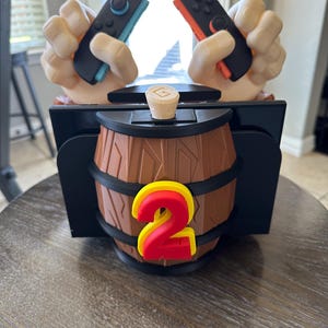 May include: A brown barrel-shaped game controller holder with a red and yellow number 2. The holder has two hands holding game controllers, one blue and black, the other red and black. A wooden stopper is on top.
