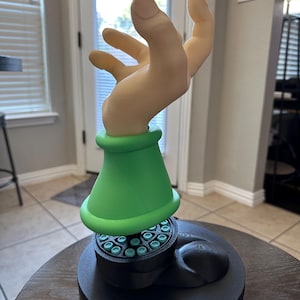 May include: A 3D-printed sculpture featuring a hand with a light skin tone, extending upwards. The hand is connected to a green conical base, which sits atop a black, circular platform with teal accents. The sculpture is on a round, wooden table.
