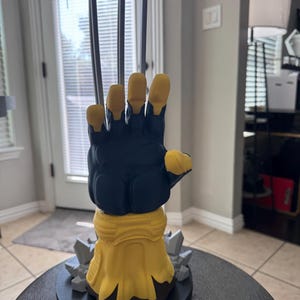 May include: A figurine of a hand with three long, sharp, metallic-colored claws extending upward. The hand is colored in dark blue and yellow, featuring a yellow wrist cuff and a base decorated with gray, jagged, rock-like elements on a black stand.