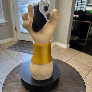May include: A decorative hand-shaped stand, holding a white and black Xbox controller. The stand has a gold-colored band around the wrist area and sits on a black circular base. The controller is a standard size.